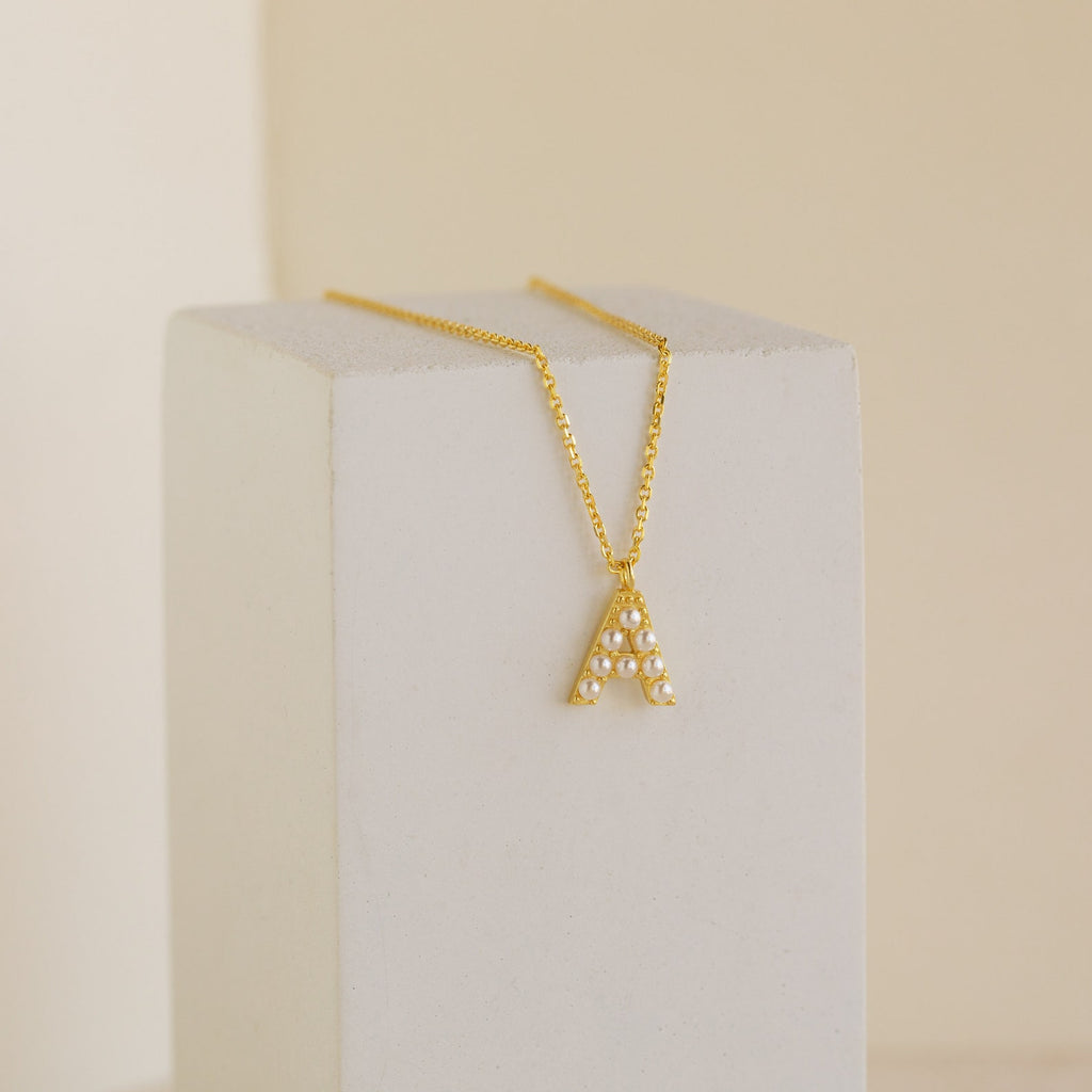 The Pearl Letter Necklace features a gold chain with a pearl-studded letter A pendant, displayed on a white block—an elegant and personalized jewelry piece that stands out in any collection.