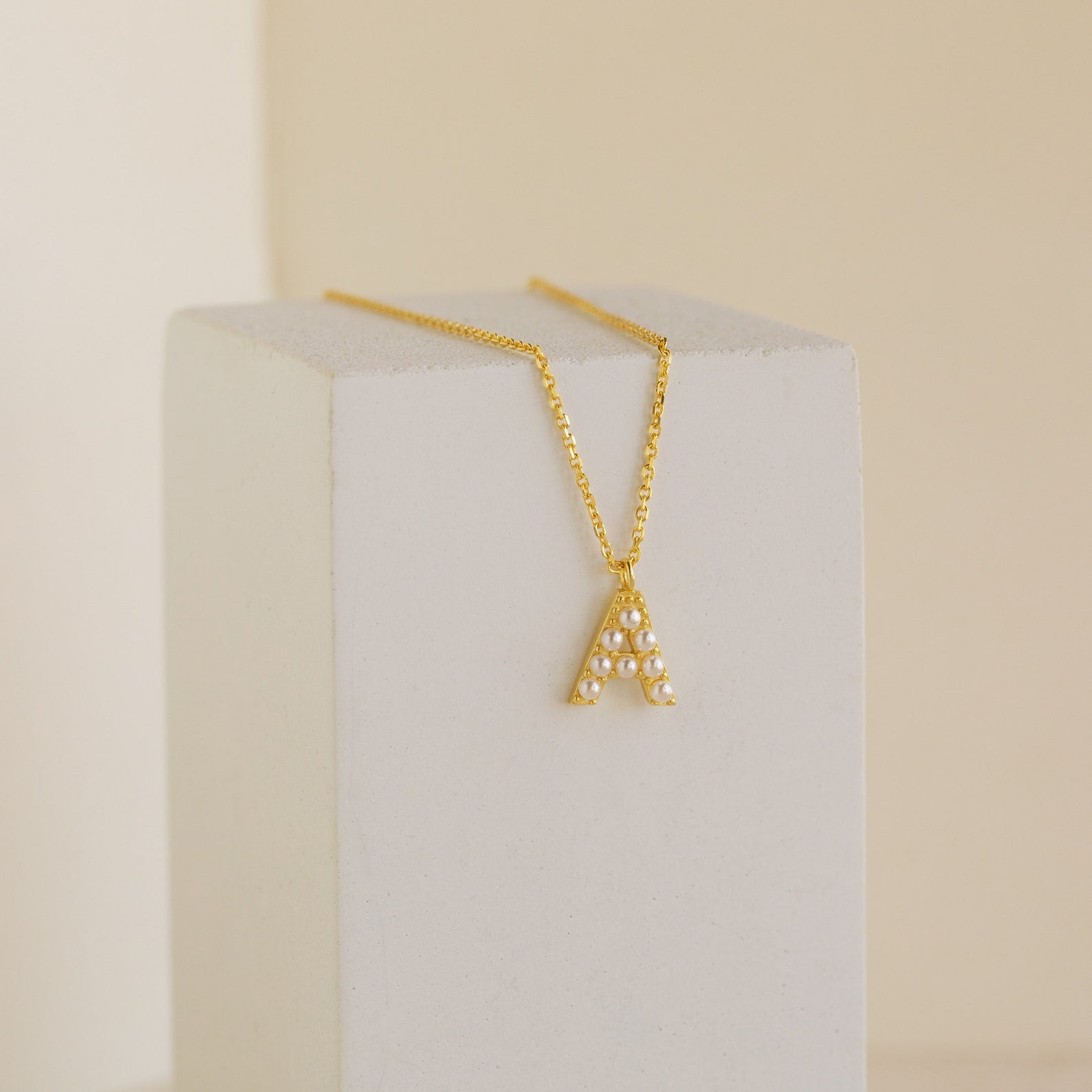 The Pearl Letter Necklace features a gold chain with a pearl-studded letter A pendant, displayed on a white block—an elegant and personalized jewelry piece that stands out in any collection.
