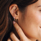 A woman smiles as she touches her ear, wearing Wren Spike Studs—small, lightweight earrings with one in silver and one in gold.
