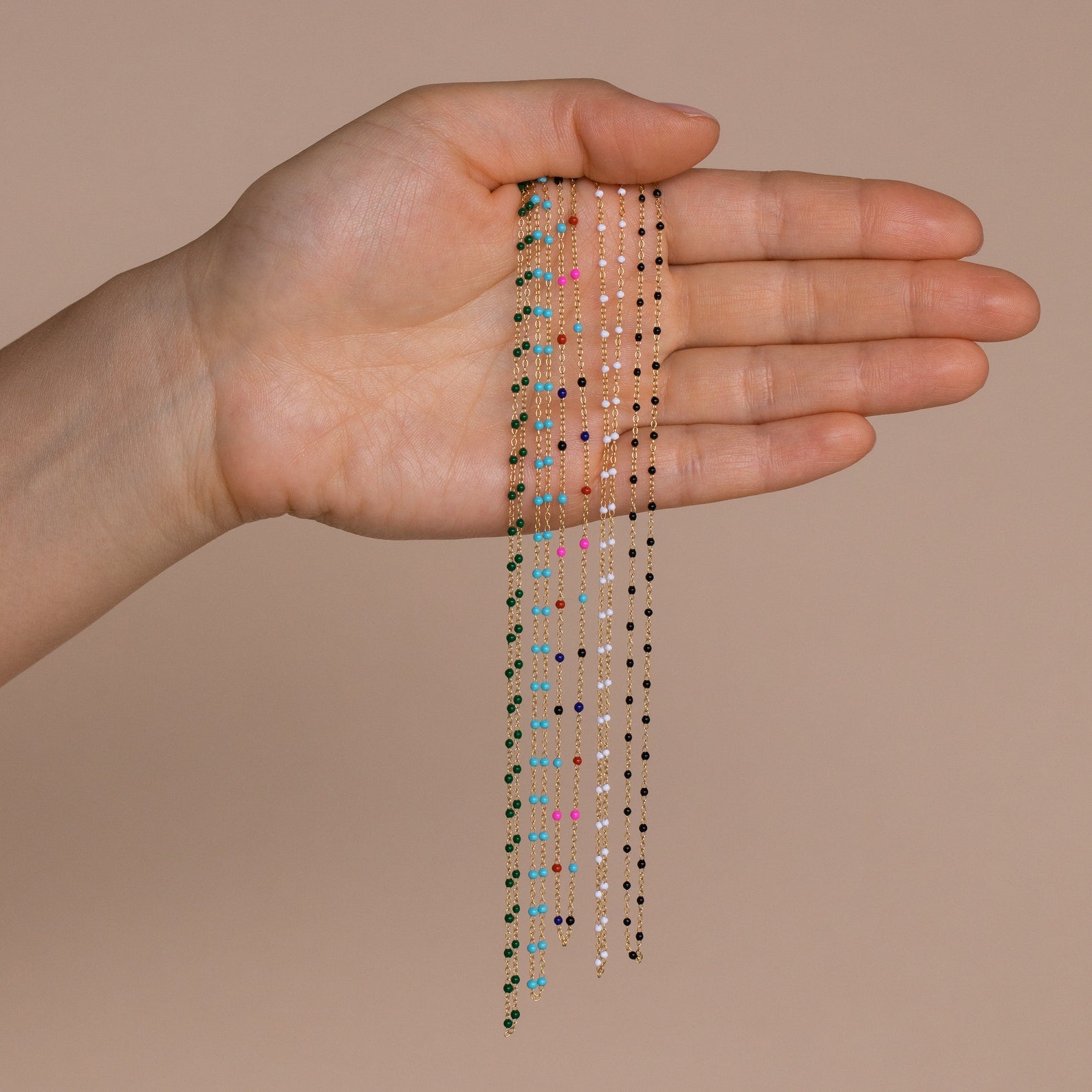 A hand displays Beaded Station Necklaces—delicate, trendy jewelry chains featuring vibrant beads—set against a neutral beige background.