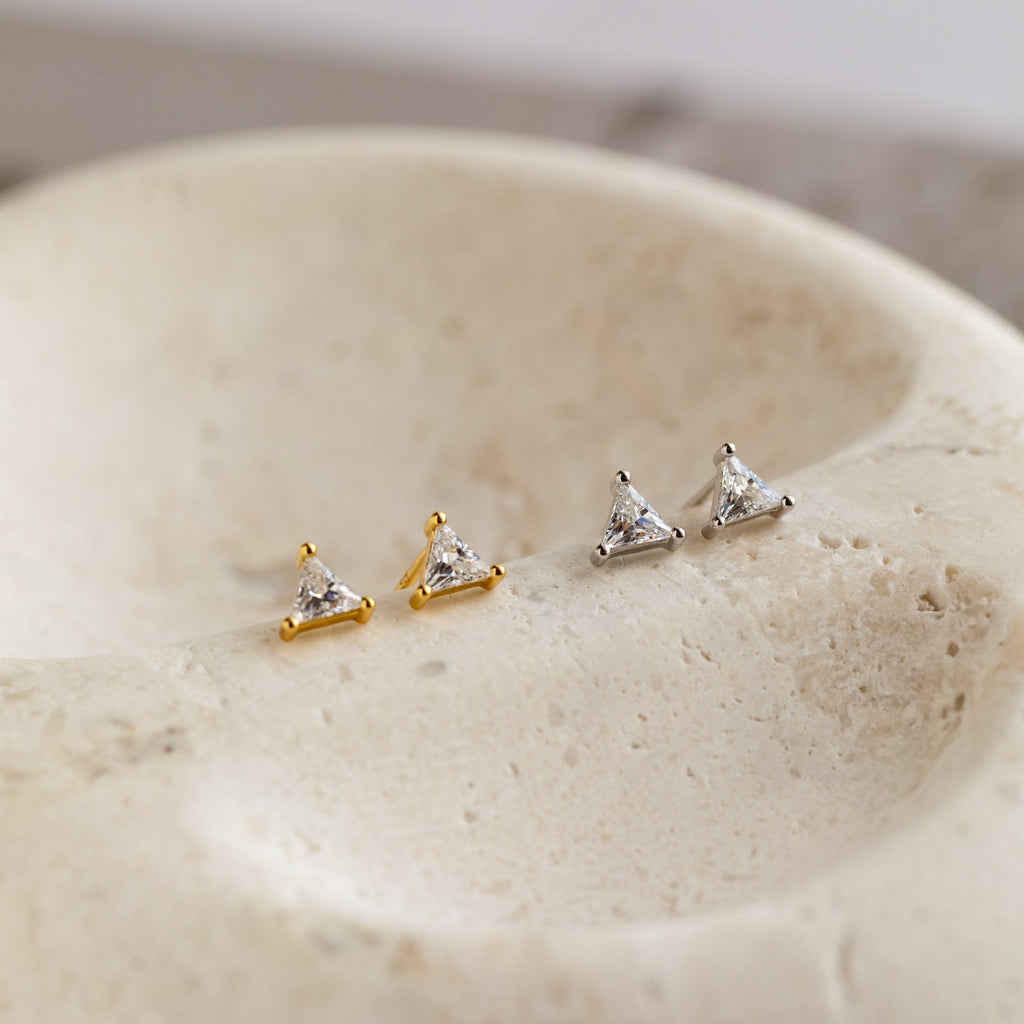 Two pairs of Trixie Diamond Studs, featuring modern triangular designs in gold and silver, are displayed on a textured stone surface.