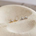 Two pairs of Trixie Diamond Studs, featuring modern triangular designs in gold and silver, are displayed on a textured stone surface.