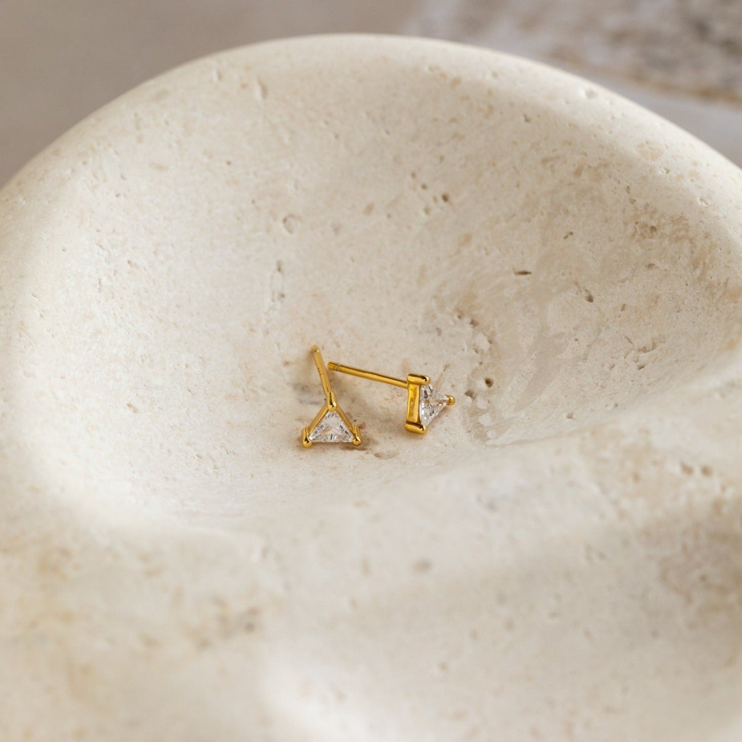 The Trixie Diamond Studs—gold, triangle-shaped earrings—rest on a textured, off-white stone surface, exemplifying modern classic jewelry style.