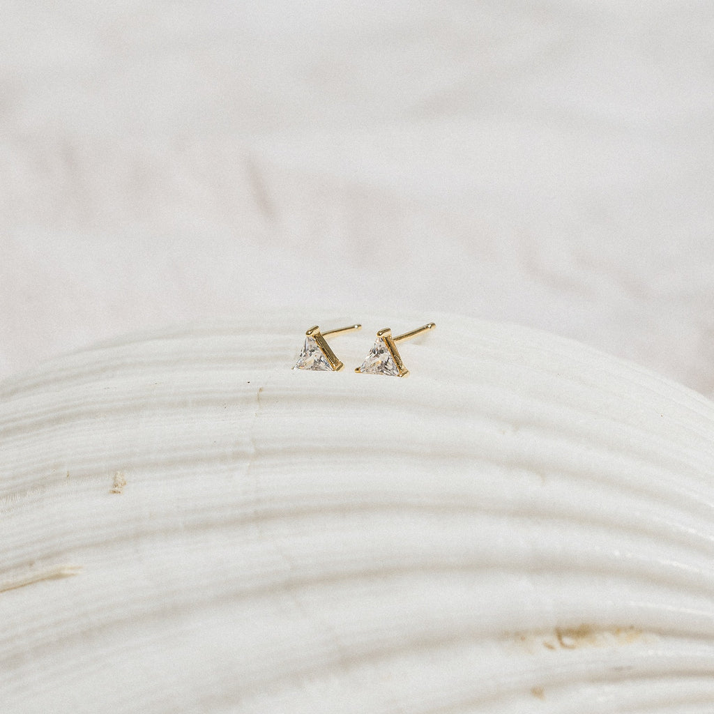The Trixie Diamond Studs, gold triangular earrings with clear stones, showcase modern classic style and are beautifully presented on a textured white seashell background.