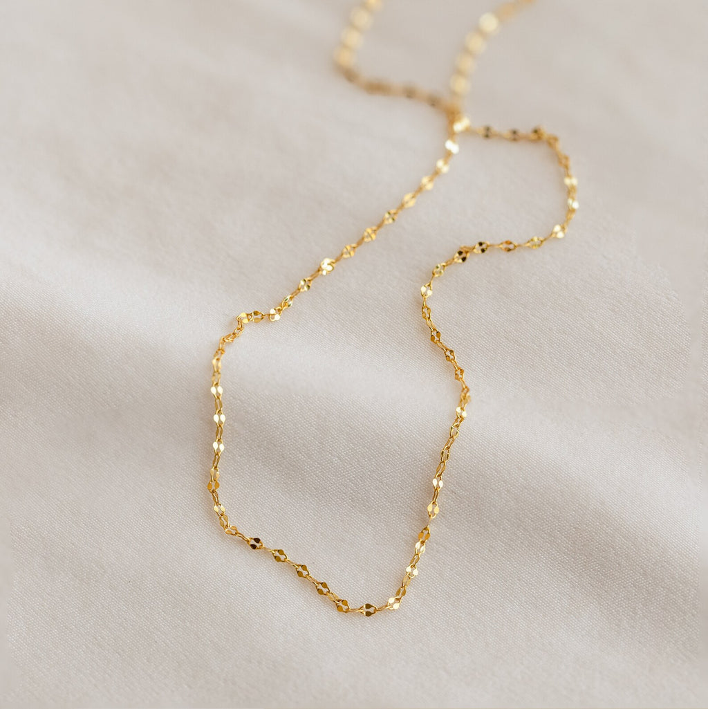 Close-up of a delicate gold chain necklace with small faceted links, displayed on a soft white fabric background.