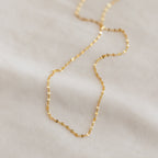 Close-up of a delicate gold chain necklace with small faceted links, displayed on a soft white fabric background.