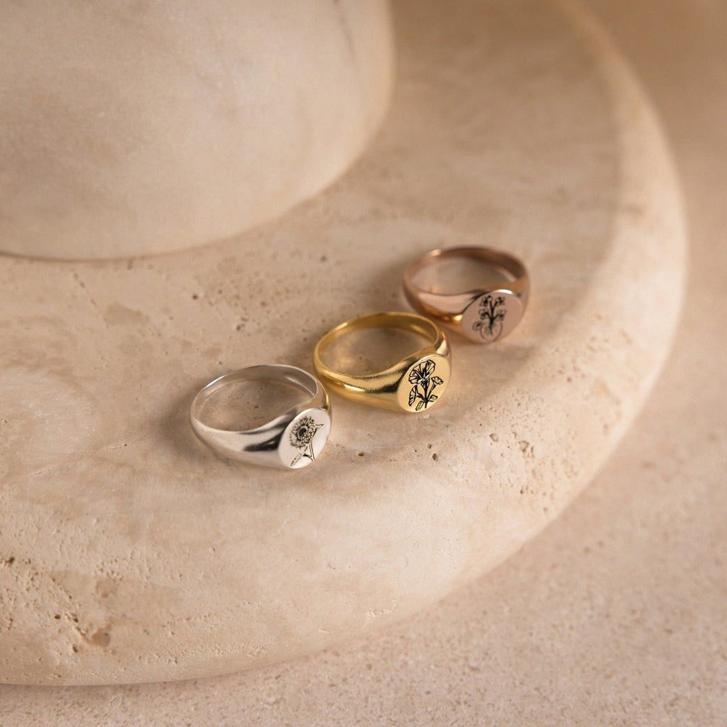 Three Birth Flower Signet Rings in silver, gold, and rose gold—ideal personalized jewelry pieces—are displayed on a round beige stone surface.
