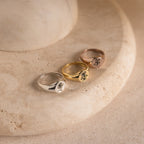 Three Birth Flower Signet Rings in silver, gold, and rose gold—ideal personalized jewelry pieces—are displayed on a round beige stone surface.