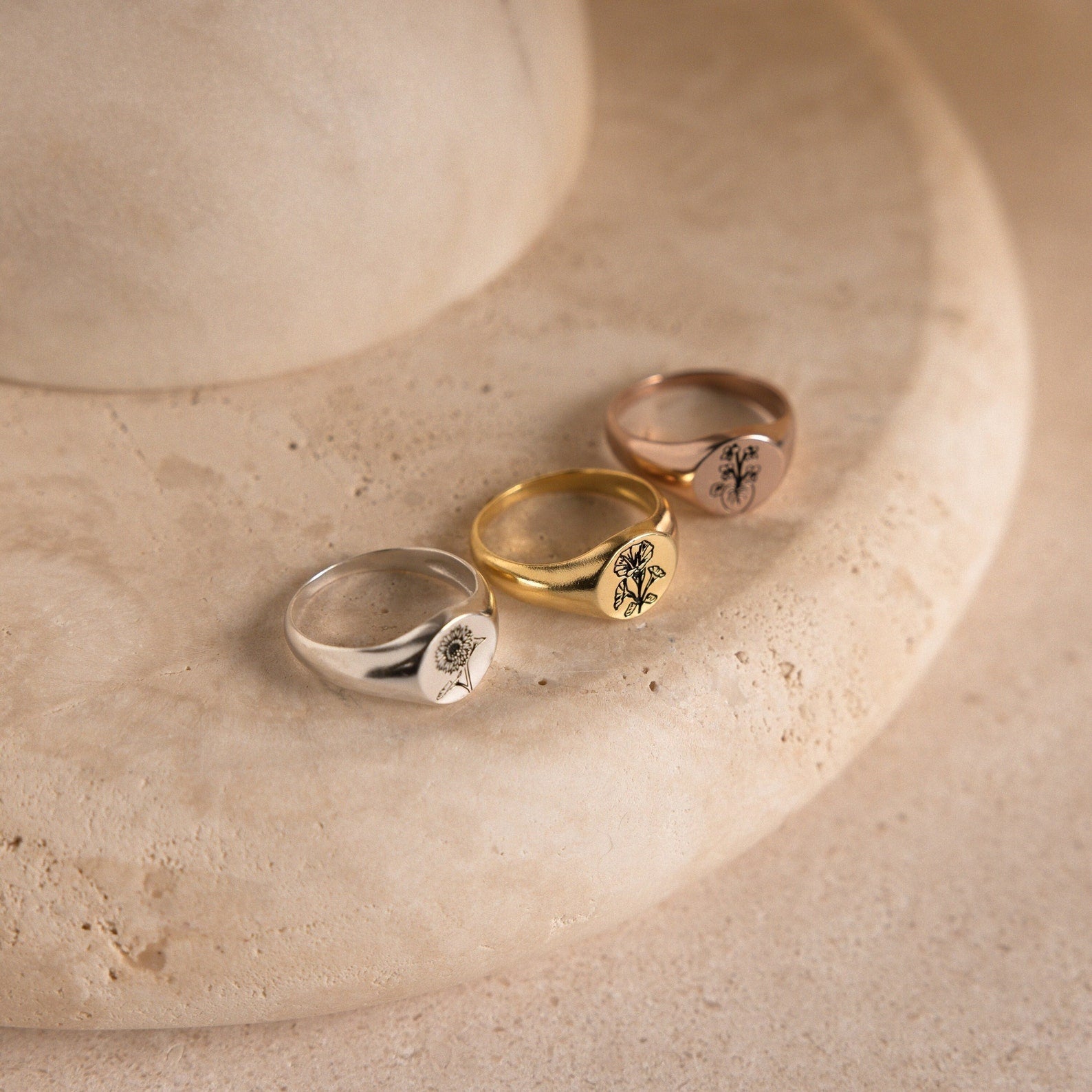 Three Birth Flower Signet Rings in silver, gold, and rose gold—ideal personalized jewelry pieces—are displayed on a round beige stone surface.