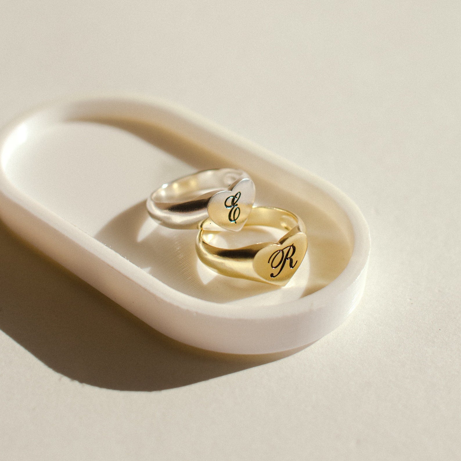 A silver and a gold Traditional Initial Heart Signet Ring rest on a white oval dish in soft natural light—an ideal anniversary gift.