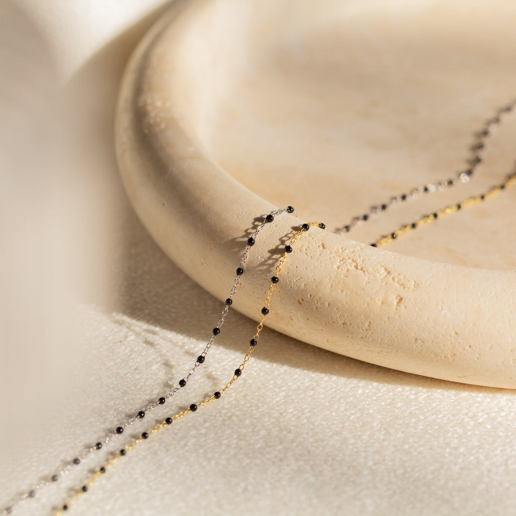 The Black Station Necklace features delicate beaded chains in gold and silver, elegantly draped over a round, light beige ceramic plate.