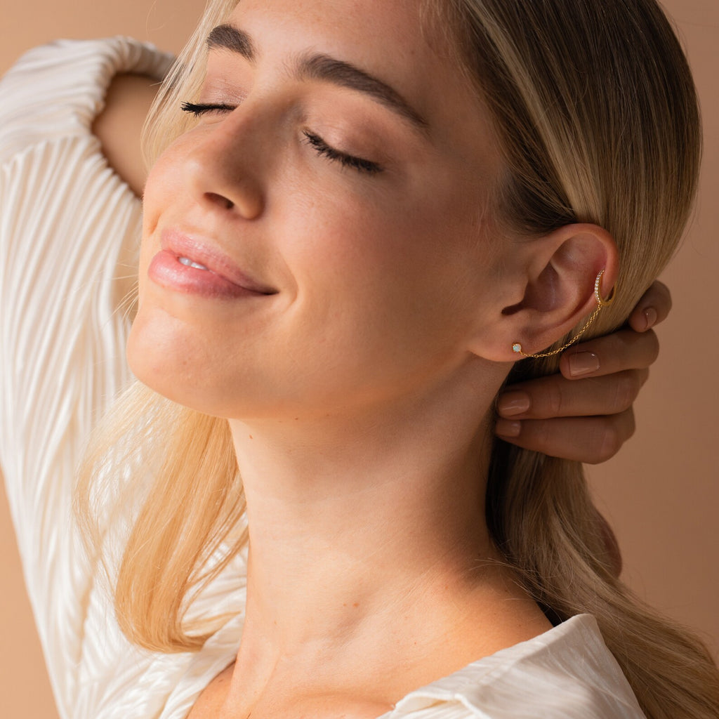Woman with closed eyes smiling softly, wearing Opal Stud & Pave Hoop Earrings and a white blouse, hand behind her head.
