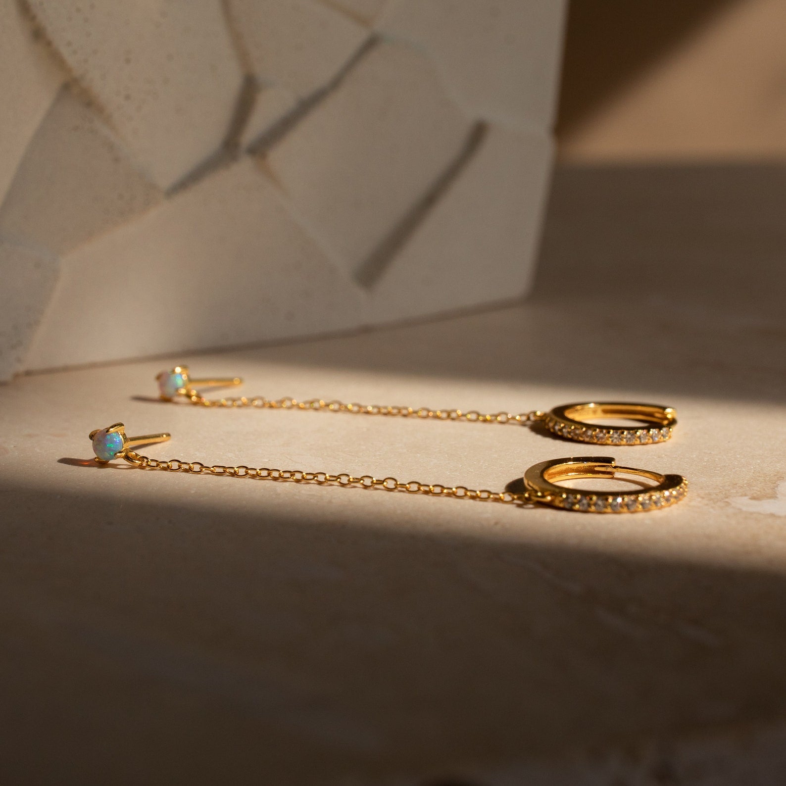 Opal Stud & Pave Hoop Earrings—gold earrings with delicate chains and gemstones—are displayed on a light surface with a geometric background.
