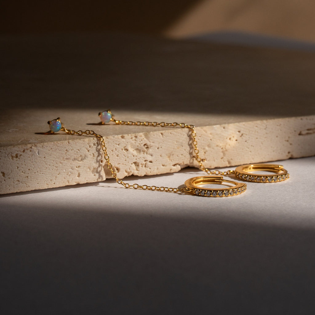 The Opal Stud & Pave Hoop Earrings, featuring gold hoops with small stones and a delicate chain, are displayed on a textured beige stone—versatile accessories perfect for any occasion.