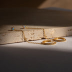 The Opal Stud & Pave Hoop Earrings, featuring gold hoops with small stones and a delicate chain, are displayed on a textured beige stone—versatile accessories perfect for any occasion.