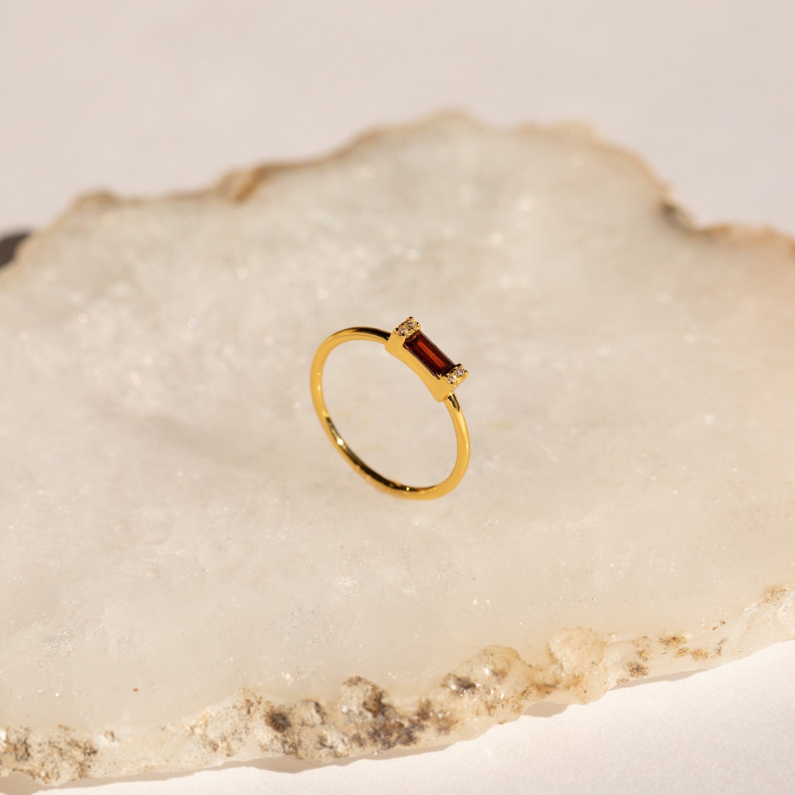 Empire Garnet Ring: a gold band ring with a rectangular deep red garnet baguette displayed on a light, textured stone surface.