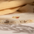 The Empire Garnet Ring features a rectangular baguette garnet set in a gold band, showcased on a textured stone surface with a beige fabric backdrop.