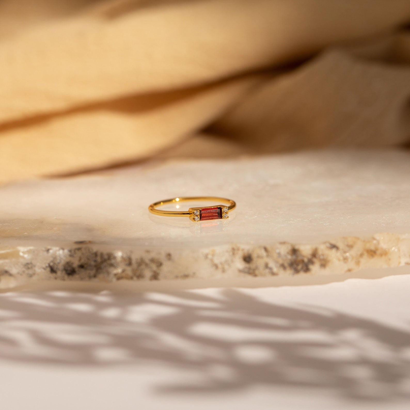 The Empire Garnet Ring features a rectangular baguette garnet set in a gold band, showcased on a textured stone surface with a beige fabric backdrop.