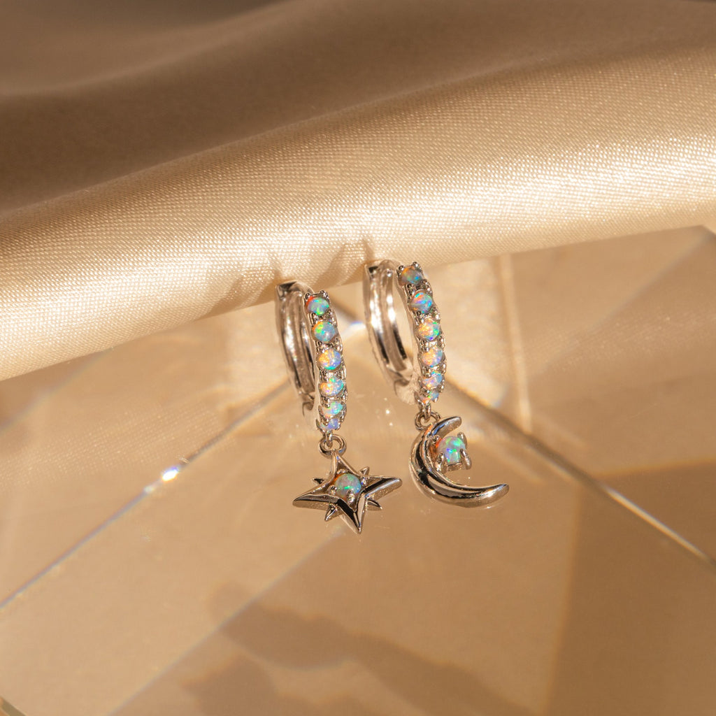 Astra Moon & Star Huggies: Silver earrings with blue gemstones—one opal-studded huggie hoop features a star charm, the other a crescent moon charm for celestial style.