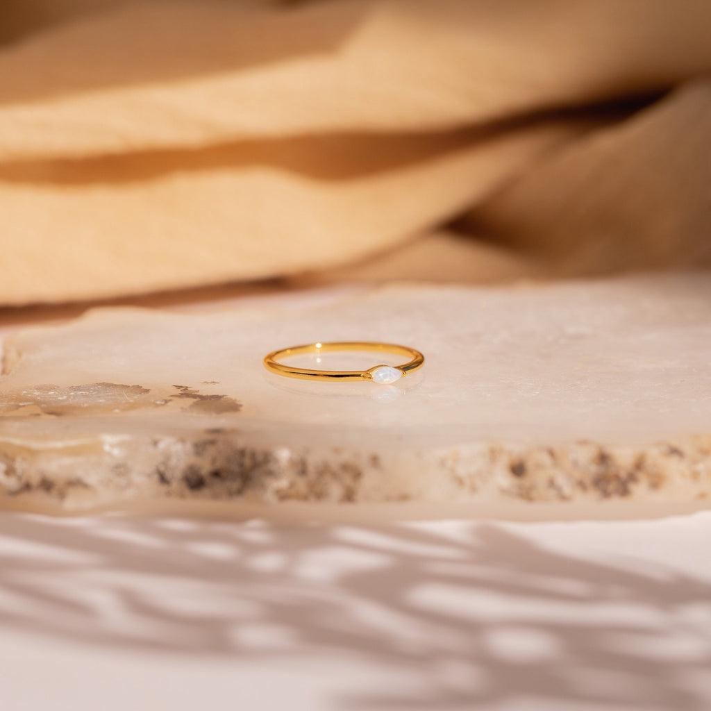 The Eloise Opal Ring, featuring a delicate gold band and a small iridescent opal gemstone, rests on a textured stone surface, embodying minimalist elegance.