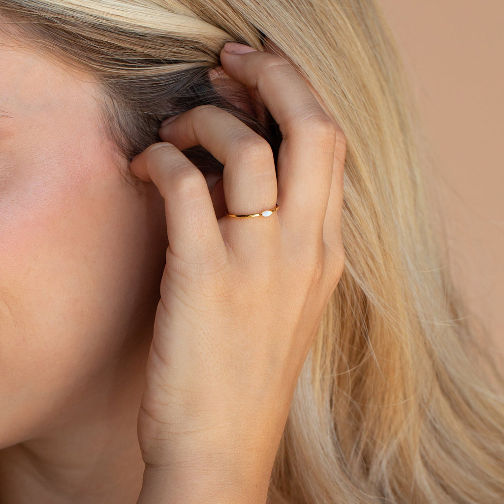 A hand wearing the Eloise Opal Ring tucks blonde hair behind an ear against a neutral background, capturing minimalist elegance.