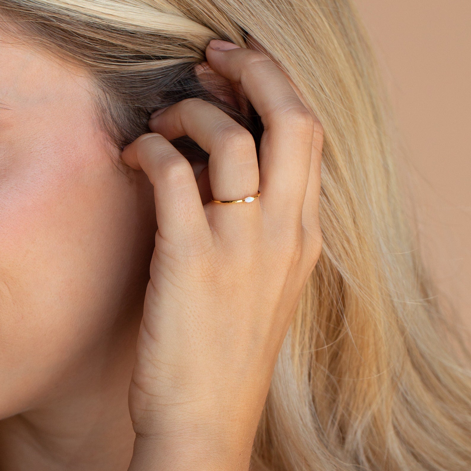 A hand wearing the Eloise Opal Ring tucks blonde hair behind an ear against a neutral background, capturing minimalist elegance.