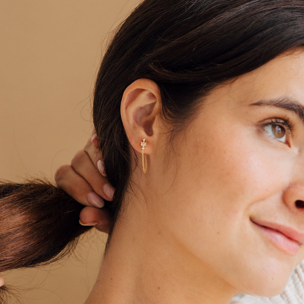 Woman with dark hair wears Florence Diamond Chain Earrings, holding her ponytail and smiling slightly against a beige background.