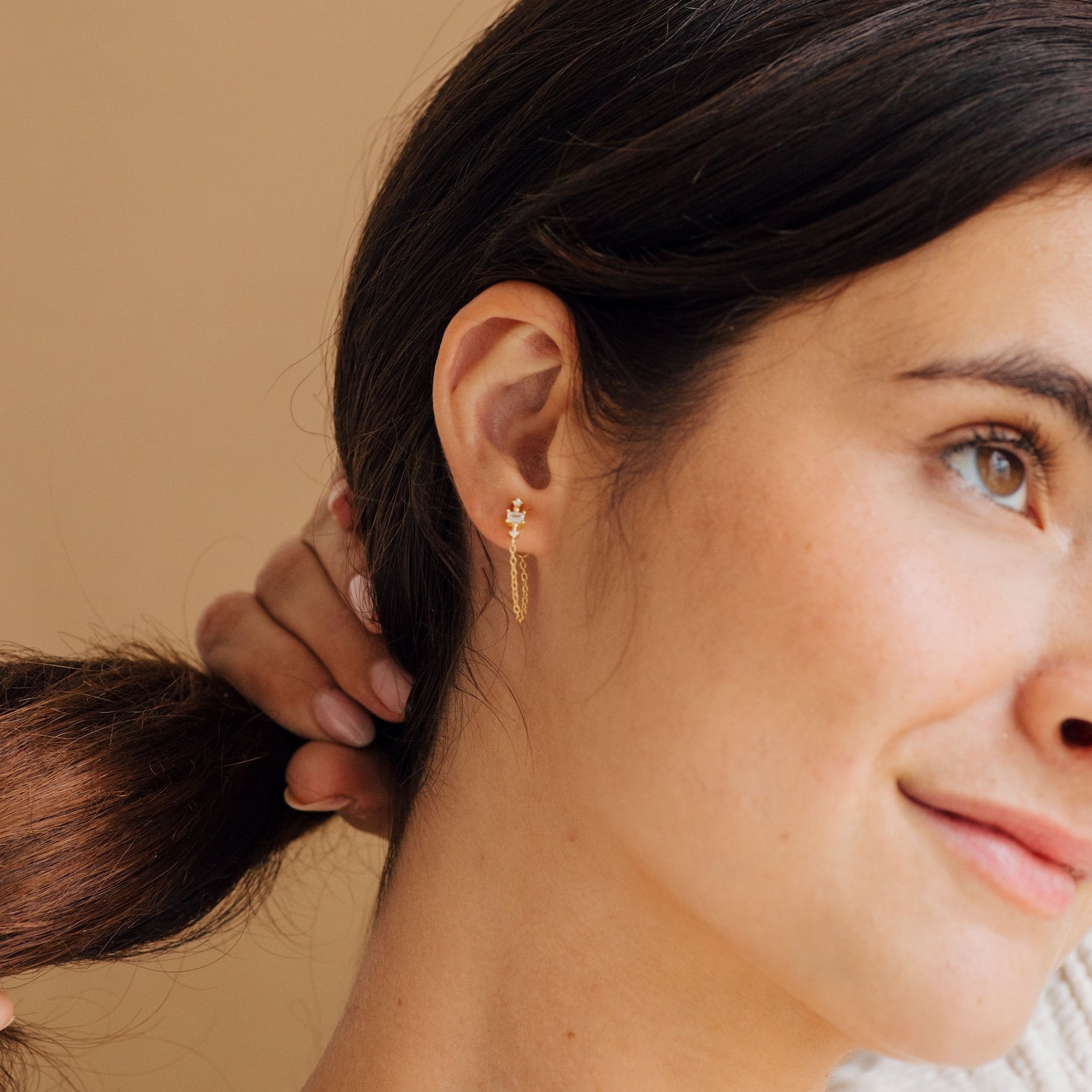 Woman with dark hair wears Florence Diamond Chain Earrings, holding her ponytail and smiling slightly against a beige background.