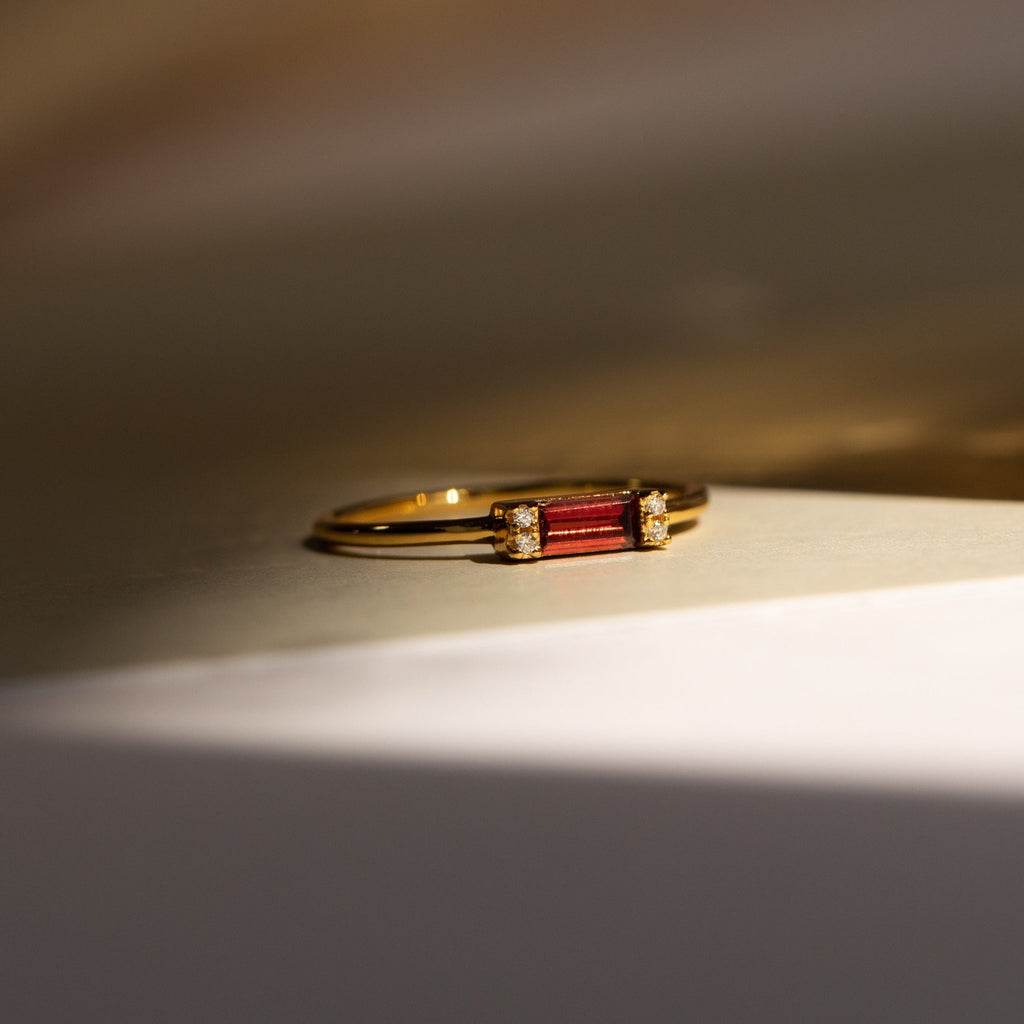 The Empire Garnet Ring features a gold band with a deep red rectangular baguette garnet and small diamonds, shown on a neutral surface in soft light.