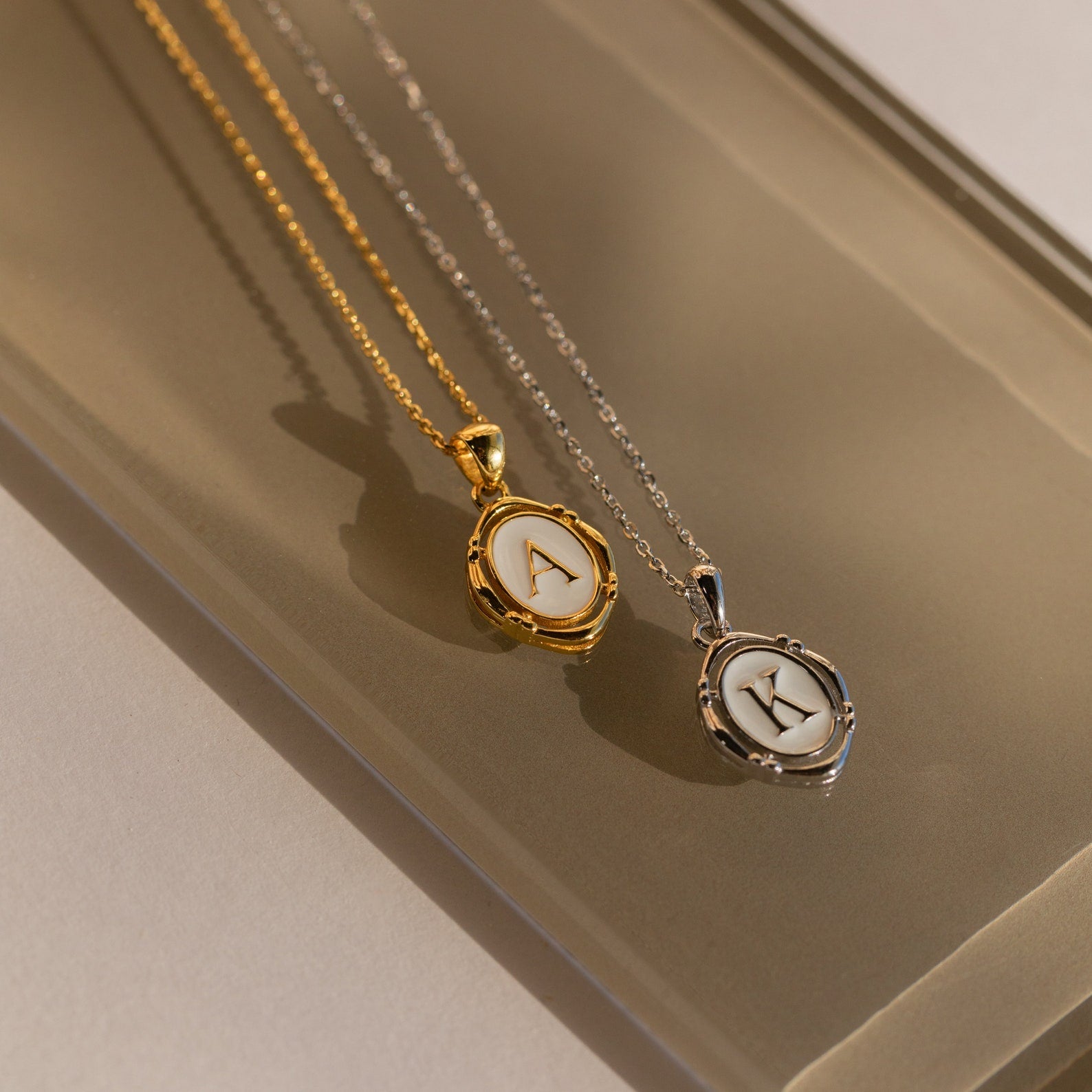 Initial Pendant Necklace in Gold with the letter A and White Gold with the letter K in a vintage serif font lay side by side.