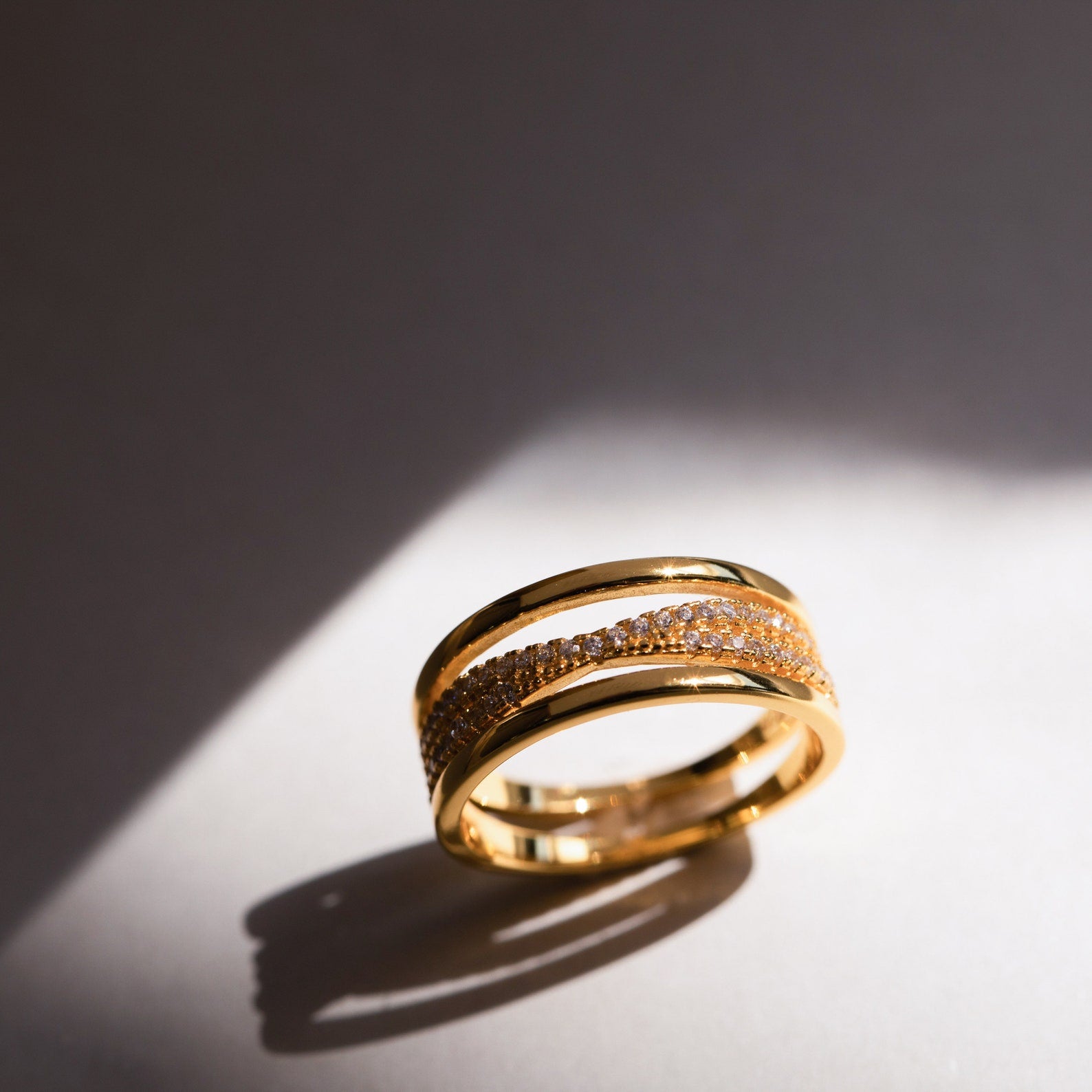 The Amani Pave Twist Ring, featuring embedded small CZ diamonds, rests on a smooth surface bathed in soft sunlight.