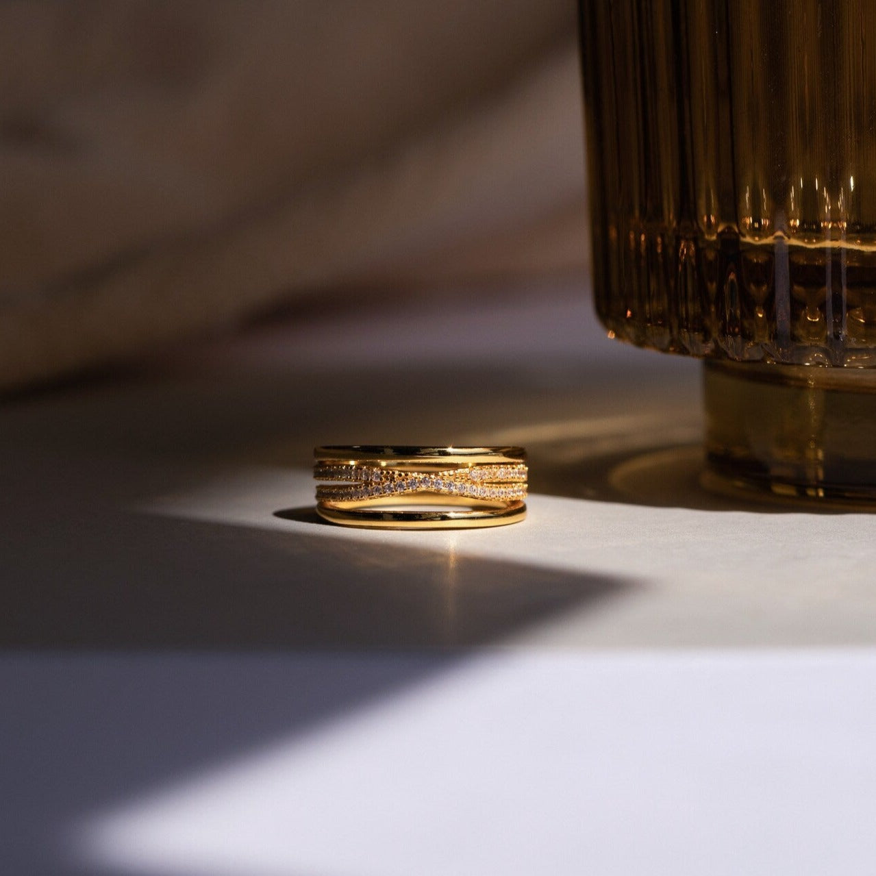 The Amani Pave Twist Ring shines with CZ diamond accents as it rests beside a textured brown glass, softly lit for a moody, elegant look.