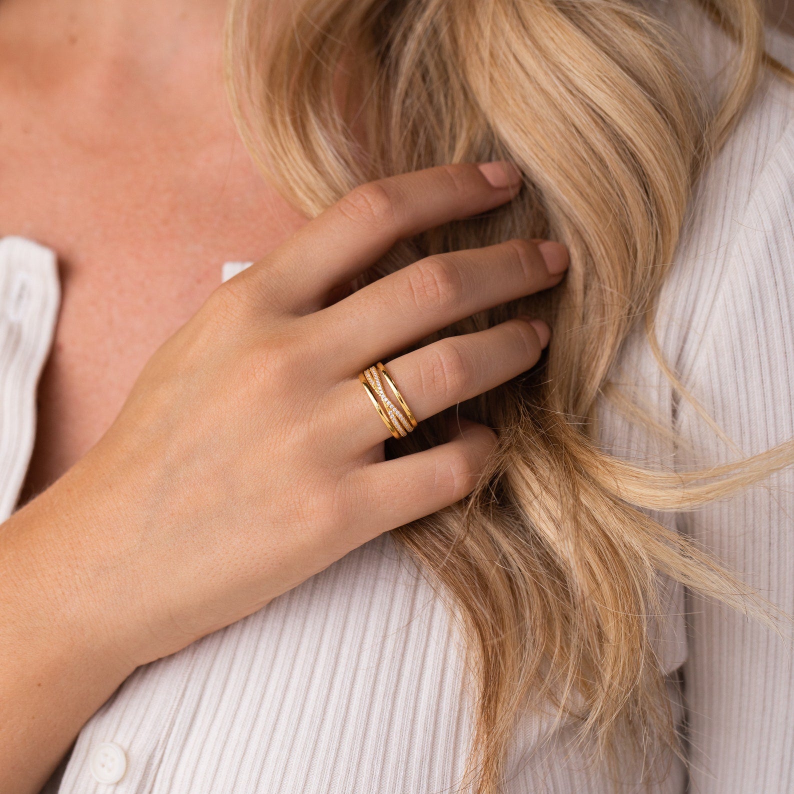 A woman with blonde hair wears stacked gold rings, including the Amani Pave Twist Ring, as she touches her hair in a white shirt.