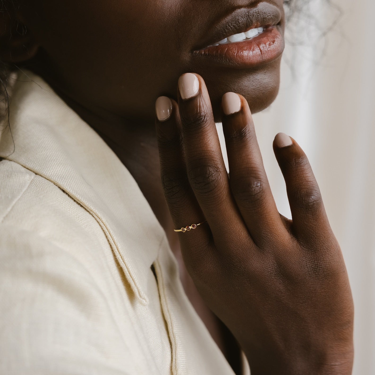 A woman with nude nail polish touches her face, showcasing the Tiny Triple Birthstone Ring with a thin band, while wearing a light-colored jacket.