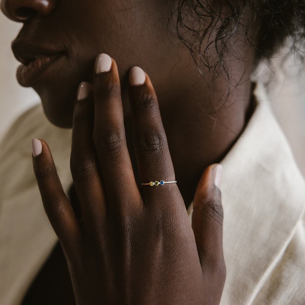 A person with light nail polish touches their face, wearing the Tiny Triple Birthstone Ring, which features a slim band set with colorful gemstones.