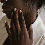A person with light nail polish touches their face, wearing the Tiny Triple Birthstone Ring, which features a slim band set with colorful gemstones.