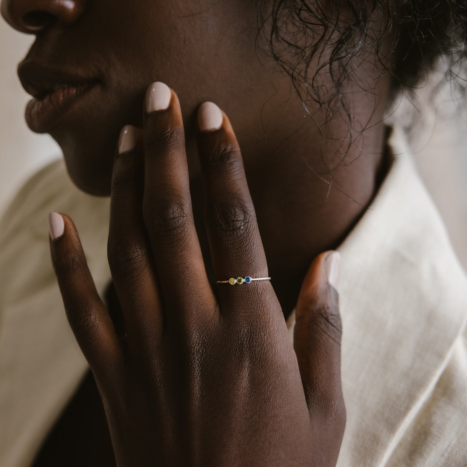 A person with light nail polish touches their face, wearing the Tiny Triple Birthstone Ring, which features a slim band set with colorful gemstones.