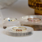Three thin band rings with colorful gemstones are displayed on round, clear stands against a soft background, showcasing the delicate style of the Tiny Triple Birthstone Ring.