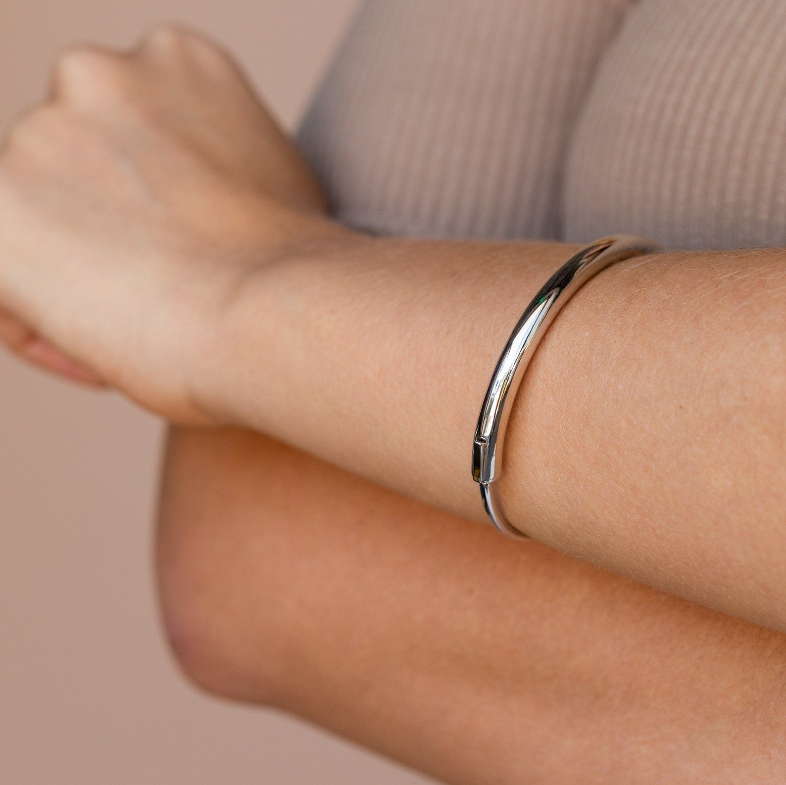 A person wearing the Aris Hinge Bangle—a simple, shiny silver cuff bracelet—on their wrist, with arms crossed.