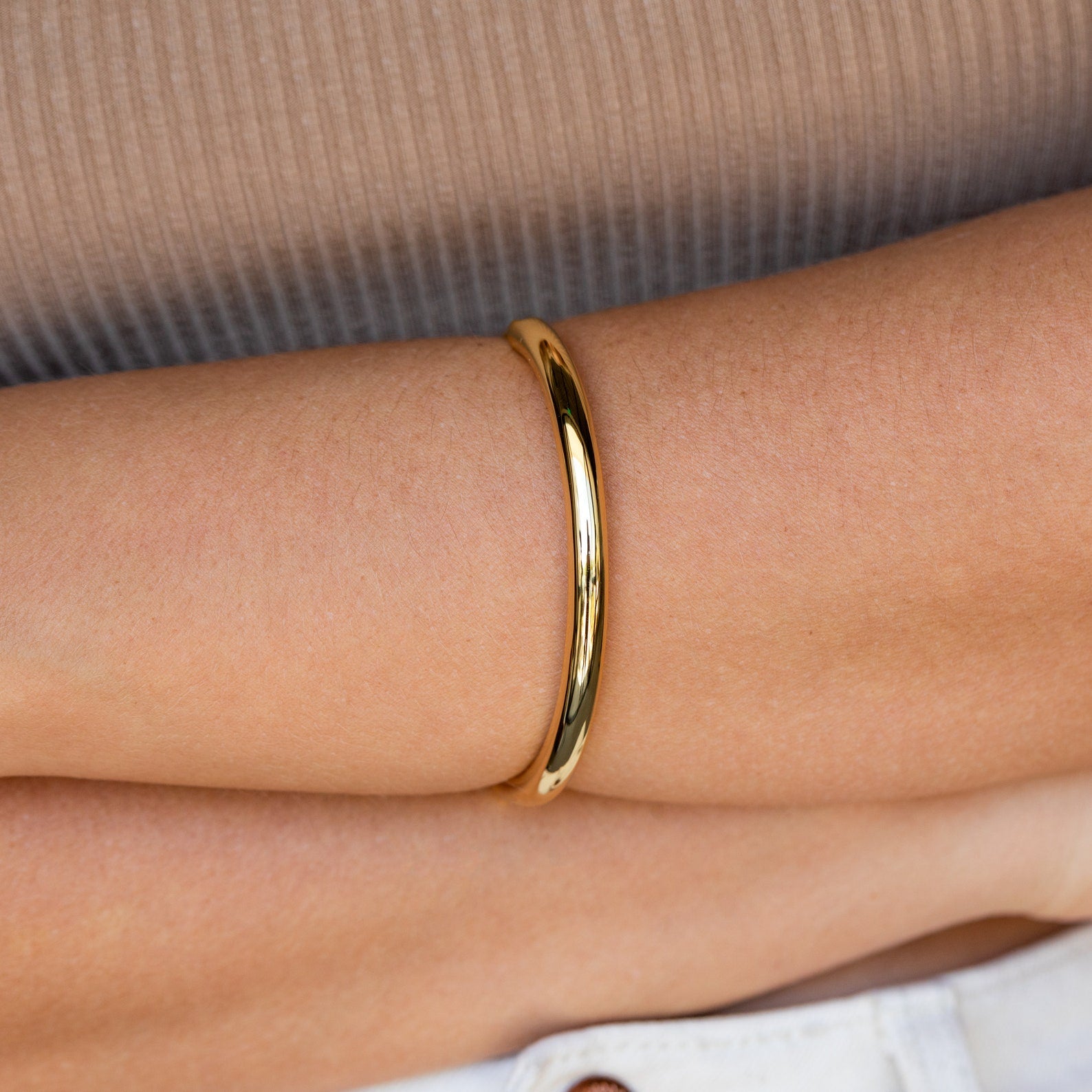 The Aris Hinge Bangle is worn on the wrist, styled with a beige ribbed shirt and light-colored pants.
