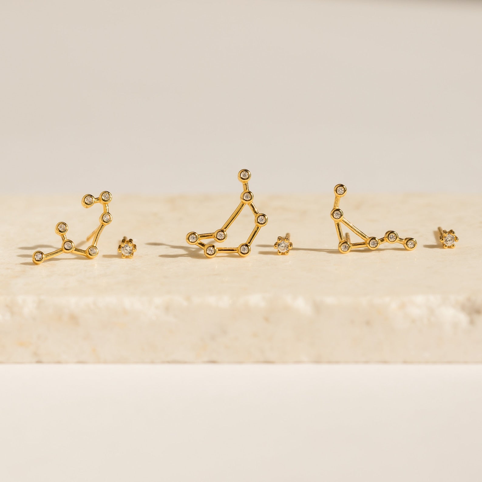 Five Constellation Earrings rest on a light stone surface, illuminated by soft lighting and inspired by astrology.