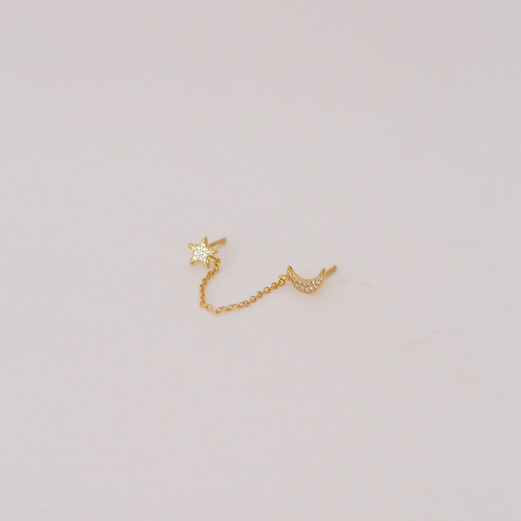 The Luna Moon & Star Chain Earrings feature gold star and moon studs linked by a delicate chain, creating unique chain stud earrings. Pictured on a simple light background.