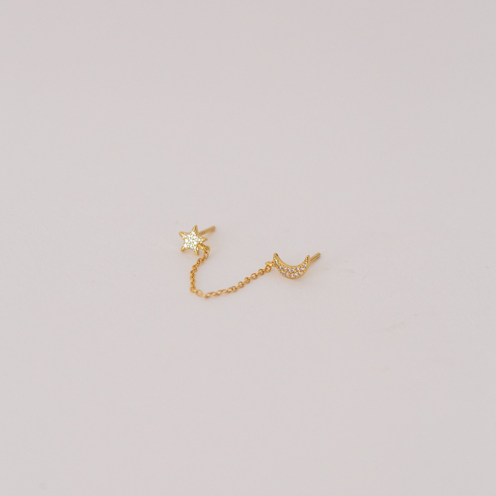 The Luna Moon & Star Chain Earrings feature gold star and moon studs linked by a delicate chain, creating unique chain stud earrings. Pictured on a simple light background.