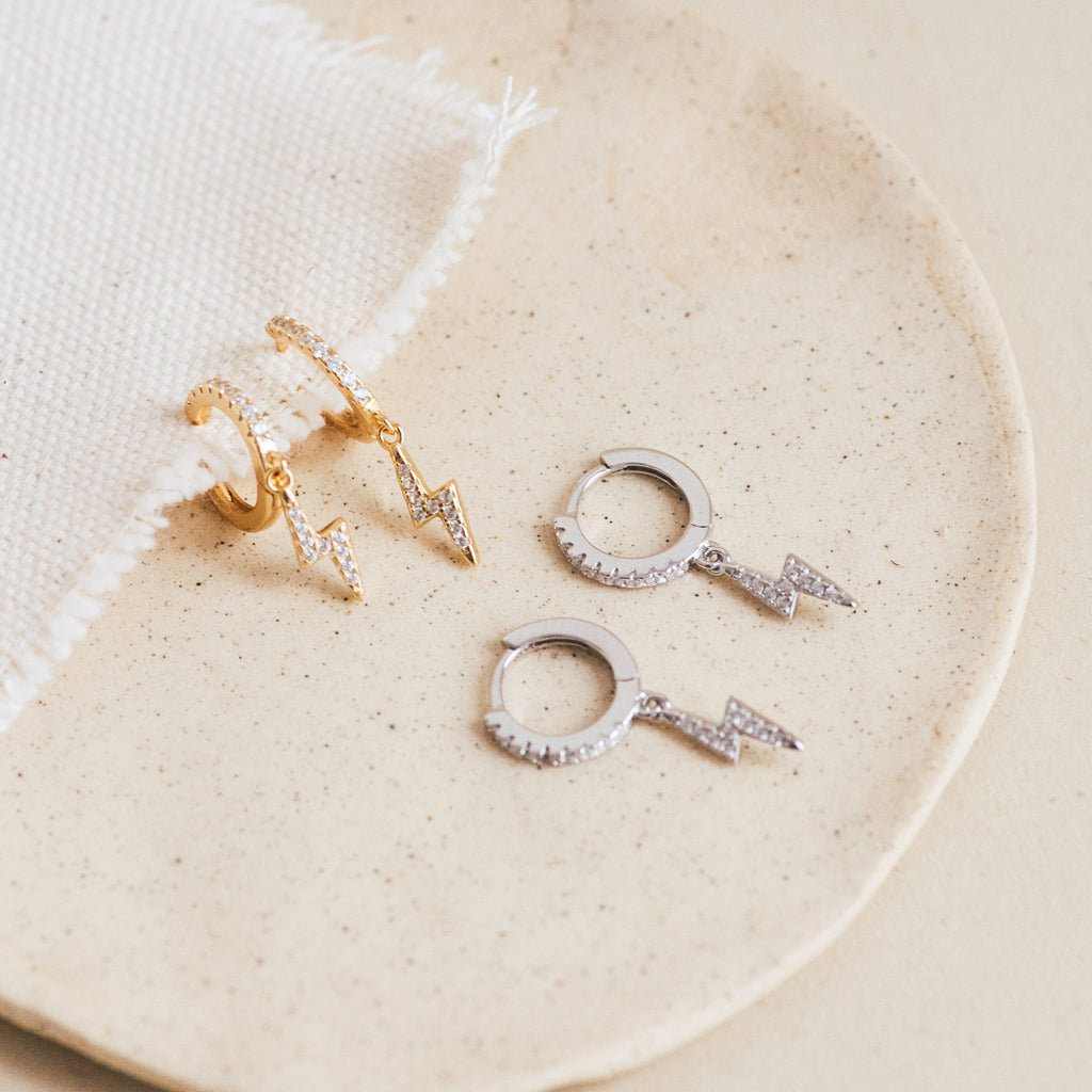Two pairs of Striker Pave Huggies—one in gold and one in silver with lightning bolt charms—rest on a beige plate, adding a bold accent to your look.