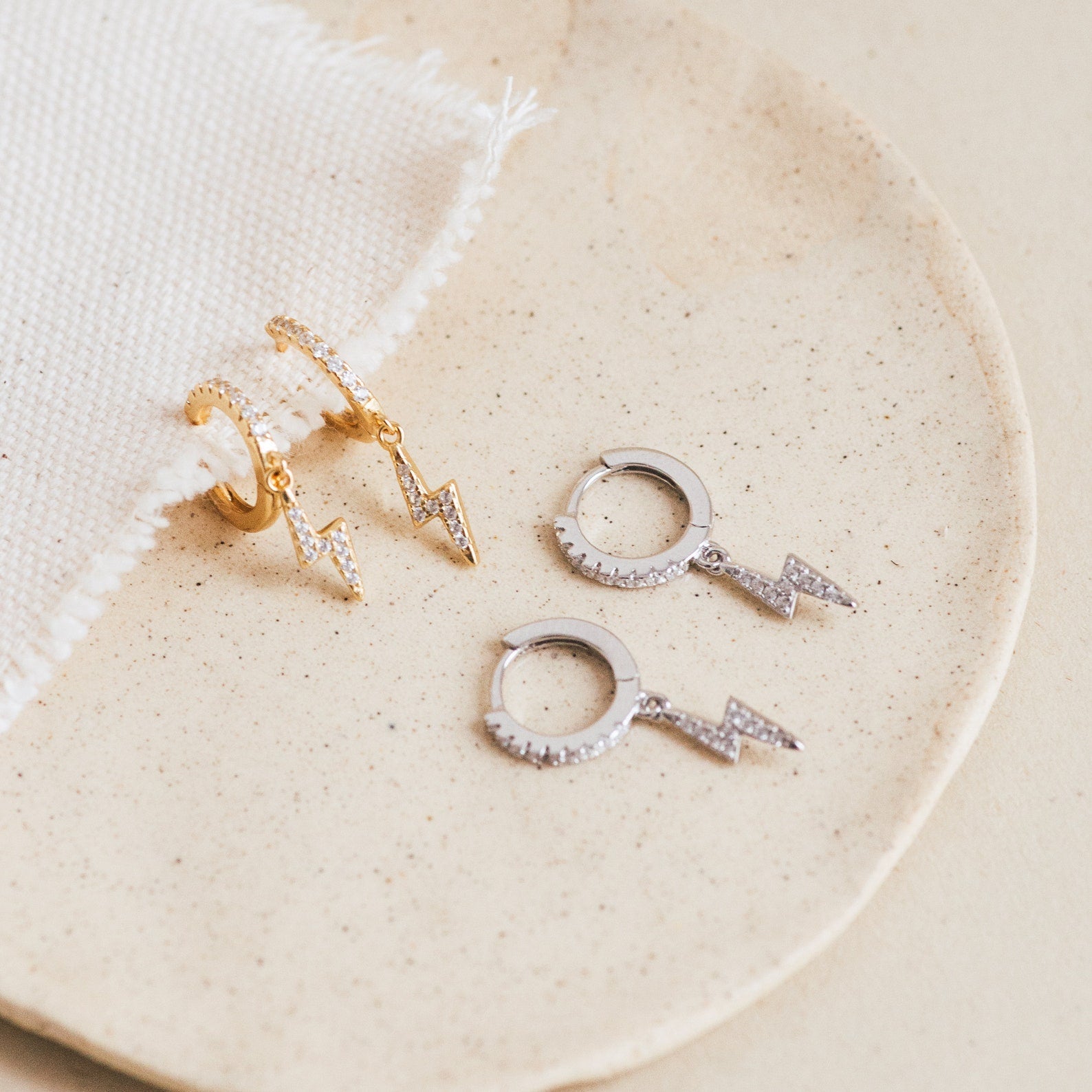 Two pairs of Striker Pave Huggies—one in gold and one in silver with lightning bolt charms—rest on a beige plate, adding a bold accent to your look.