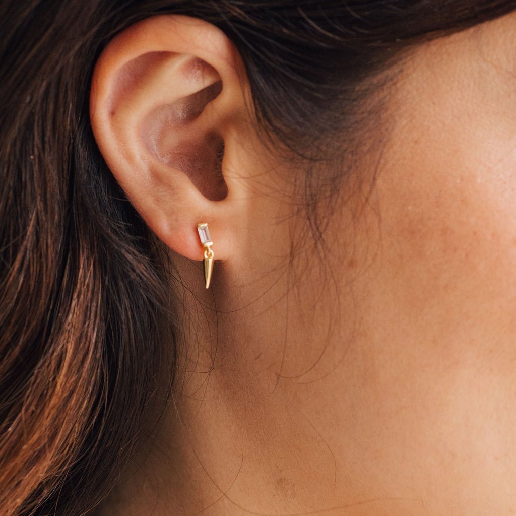 Close-up of a woman's ear wearing the Wren Spike Studs—small gold earrings with a clear CZ baguette diamond and a pointed spike drop charm in a lightweight design.