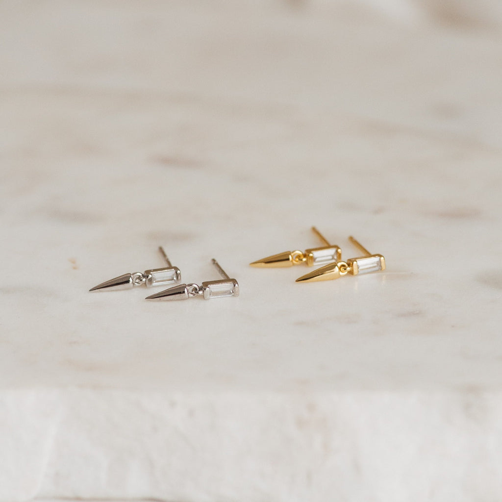 Two pairs of Wren Spike Studs, featuring spike-shaped designs in silver and gold with CZ baguette diamonds, rest elegantly on a marble surface.