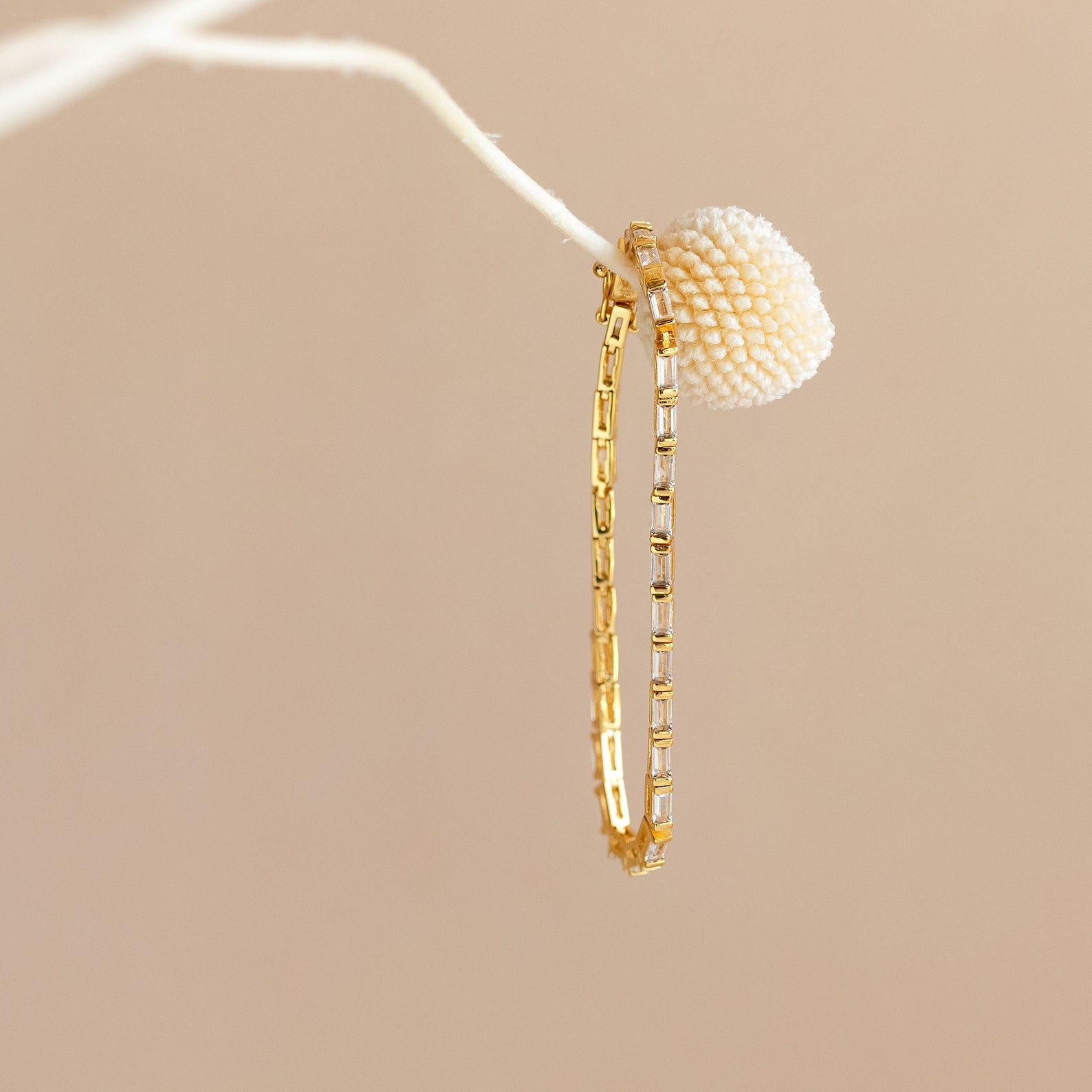 The Claudia Baguette Tennis Bracelet draped gracefully on a white branch with a textured cream bud, capturing the refined allure of elegant jewelry.