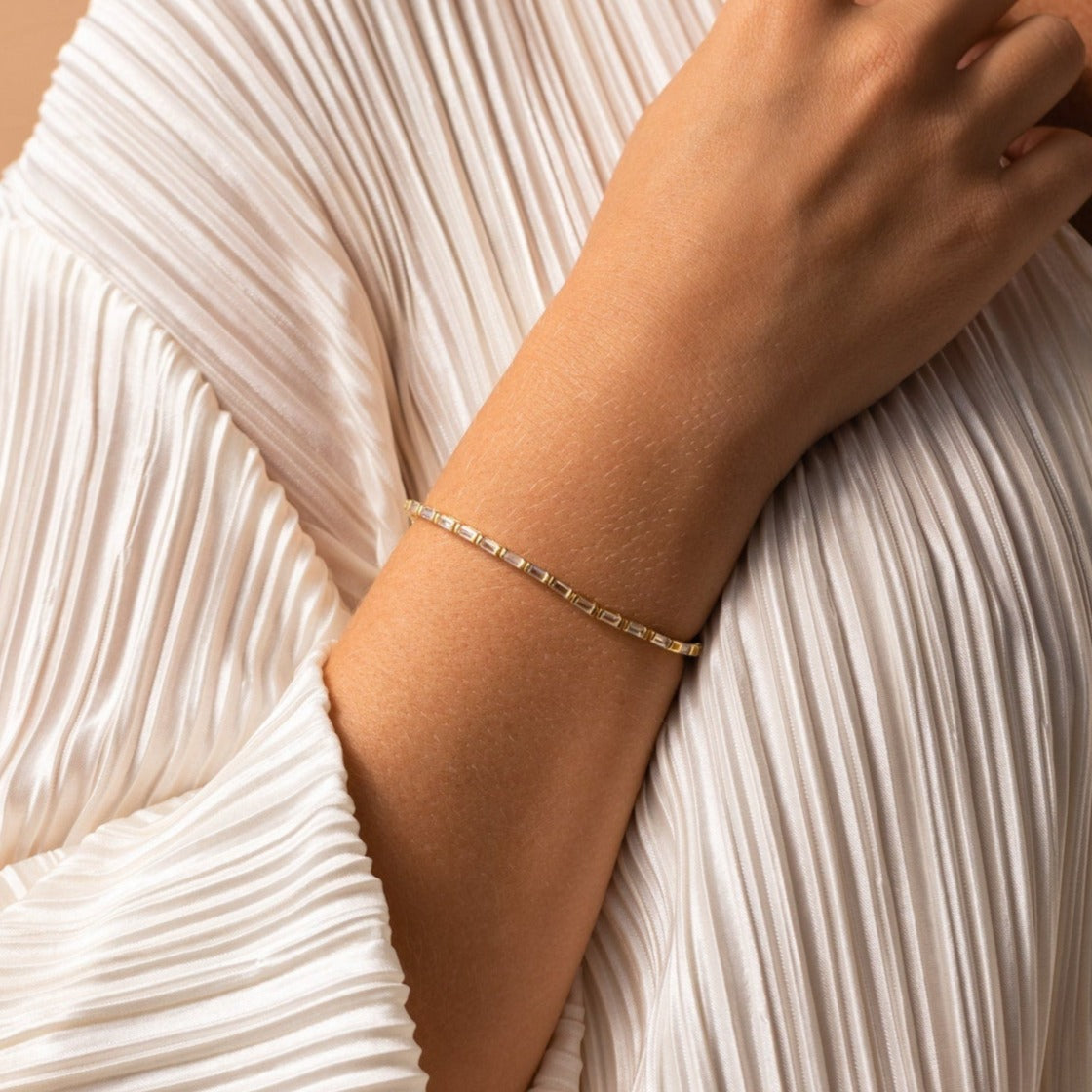 The Claudia Baguette Tennis Bracelet, featuring gold and sparkling baguette diamonds, adorns the wrist elegantly as it rests on white pleated fabric.