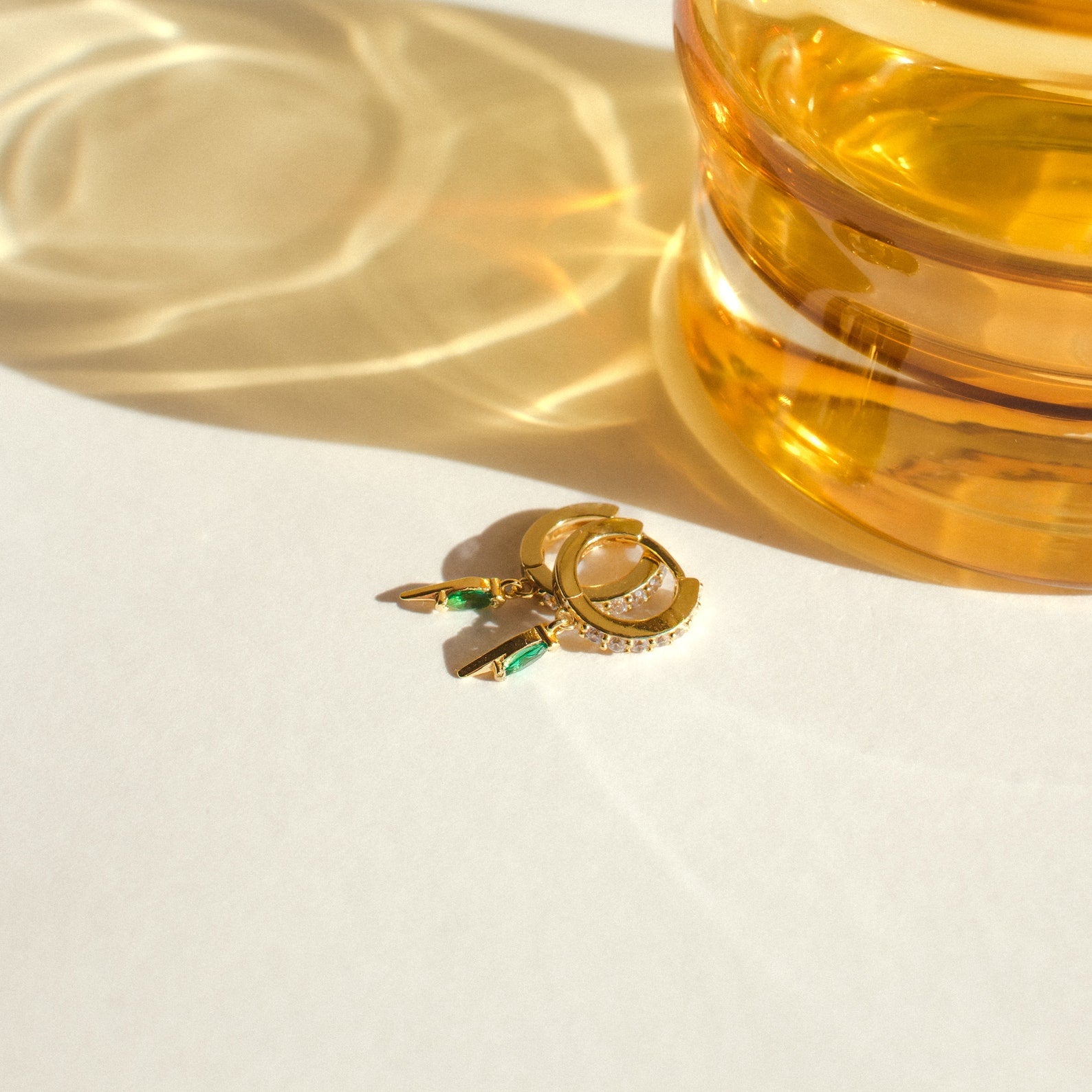 The Raine Emerald Dagger Huggies, gold hoop earrings with green gemstones, rest next to an amber glass casting reflections on a white surface.
